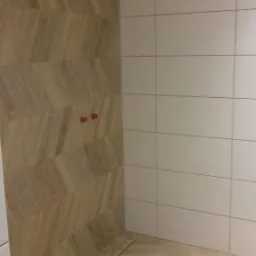 Wykończona łazienka z beżowymi płytkami ułożonymi w jodełkę na podłodze i ścianie, kontrastującymi z białymi, prostokątnymi płytkami na ścianie. Widoczny liniowy odpływ pod prysznicem i wystające...