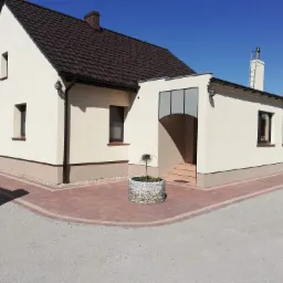 Ocieplony dom jednorodzinny z brązowym dachem i elewacją w odcieniach beżu, z widocznym oknem, kominem i małym drzewkiem w donicy na wybrukowanym podjeździe.