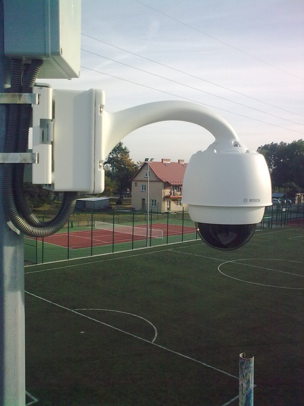 Kamera kopułkowa Bosch na słupie, skierowana na boisko sportowe z budynkiem w tle.