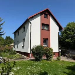 Dwukondygnacyjny dom z biało-czerwoną elewacją, widoczny z ogrodu pełnego kwiatów pod błękitnym niebem.