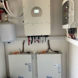 Smart Energy Fotowoltaika Paweł Szczotka - Kompletna instalacja: falownik Deye z ekranem, dwa magazyny energii Deye, zabezpieczenia i zbiornik wyrównawczy. Elementy połączone przewodami w białym pomieszczeniu.