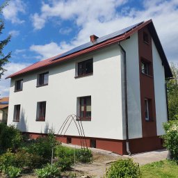 Smart Energy Fotowoltaika Paweł Szczotka - Dwukondygnacyjny dom z panelami słonecznymi na czerwonym dachu w Milówce. Biała elewacja z brązowymi akcentami. Widok z ogrodu w słoneczny dzień.