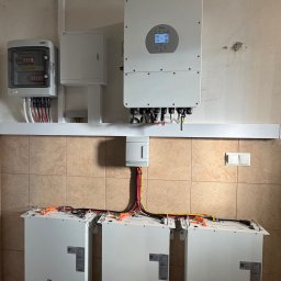 Smart Energy Fotowoltaika Paweł Szczotka - Instalacja magazynu energii Deye: trzy białe moduły na ścianie z brązowymi płytkami, inwerter i rozdzielnia elektryczna. Widoczne przewody i złącza.