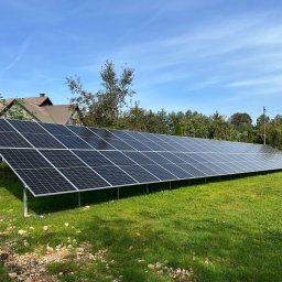 Smart Energy Fotowoltaika Paweł Szczotka - Długi rząd paneli fotowoltaicznych na zielonej trawie, z domem w tle pod błękitnym niebem. Instalacja na gruncie, widoczne mocowania i kable.
