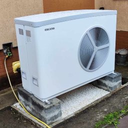 Smart Energy Fotowoltaika Paweł Szczotka - Biała pompa ciepła Stiebel Eltron na betonowych blokach i białym żwirze przed budynkiem z oknami, widoczny fragment żółtego węża.