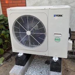 Smart Energy Fotowoltaika Paweł Szczotka - Biała jednostka zewnętrzna pompy ciepła YORK, umieszczona na betonowym fundamencie z szarym żwirem, na tle ściany budynku z cegły i betonu.