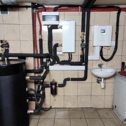 Smart Energy Fotowoltaika Paweł Szczotka - Złożona instalacja grzewcza w kotłowni: zbiorniki, rury z izolacją, sterownik. Widoczne elementy instalacji wodnej i kanalizacyjnej. Tło stanowią kafelki.