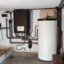 Smart Energy Fotowoltaika Paweł Szczotka - Zestaw kotłowni w piwnicy: czarny kocioł z wyświetlaczem, biały zbiornik akumulacyjny, białe naczynie przeponowe, orurowanie. Widoczne liczniki i zawory.