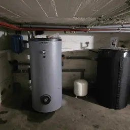 Szary zbiornik Trinnity z manometrem i czarny zbiornik w piwnicy, widoczne rury i małe zbiorniki ciśnieniowe. Instalacja w surowym wnętrzu.