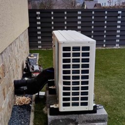 Smart Energy Fotowoltaika Paweł Szczotka - Biała pompa ciepła na betonowej podstawie przy ścianie domu. Widoczne czarne rury, trawnik i puszka po napoju. W tle ogrodzenie i budynki.