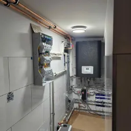 Węzeł cieplny w kotłowni: czarny zasobnik, rozdzielnia elektryczna z okablowaniem na kafelkowanej ścianie, rury miedziane pod sufitem, pompa obiegowa Wilo.
