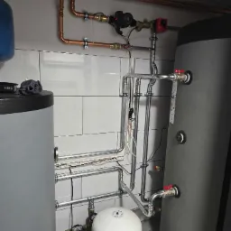 Instalacja rur miedzianych i stalowych z zaworami, manometrami i pompą obiegową w kotłowni z dwoma zbiornikami akumulacyjnymi, na tle białych płytek.