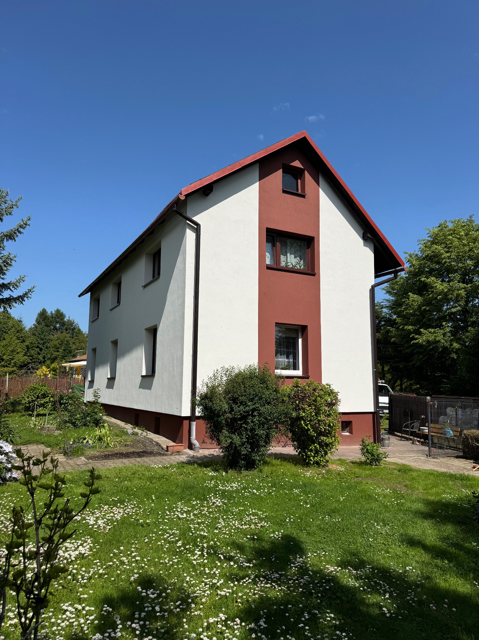 Dwukondygnacyjny dom z biało-czerwoną elewacją, widoczny z ogrodu pełnego kwiatów pod błękitnym niebem.