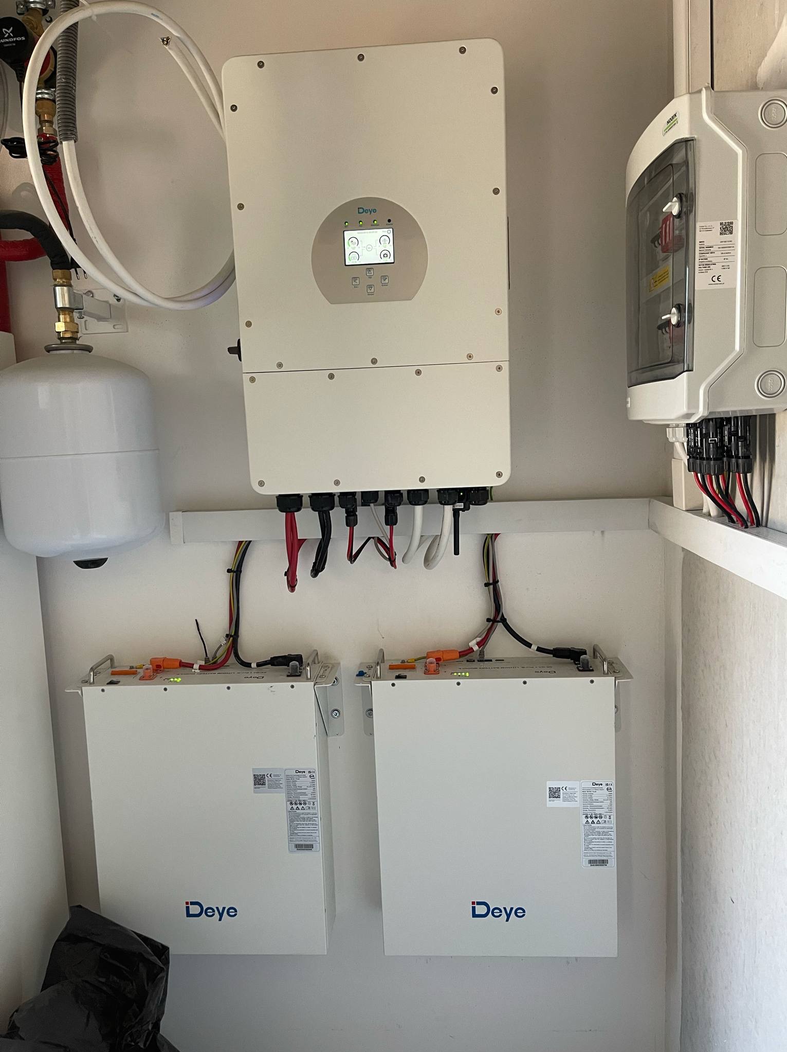 Kompletna instalacja: falownik Deye z ekranem, dwa magazyny energii Deye, zabezpieczenia i zbiornik wyrównawczy. Elementy połączone przewodami w białym pomieszczeniu.