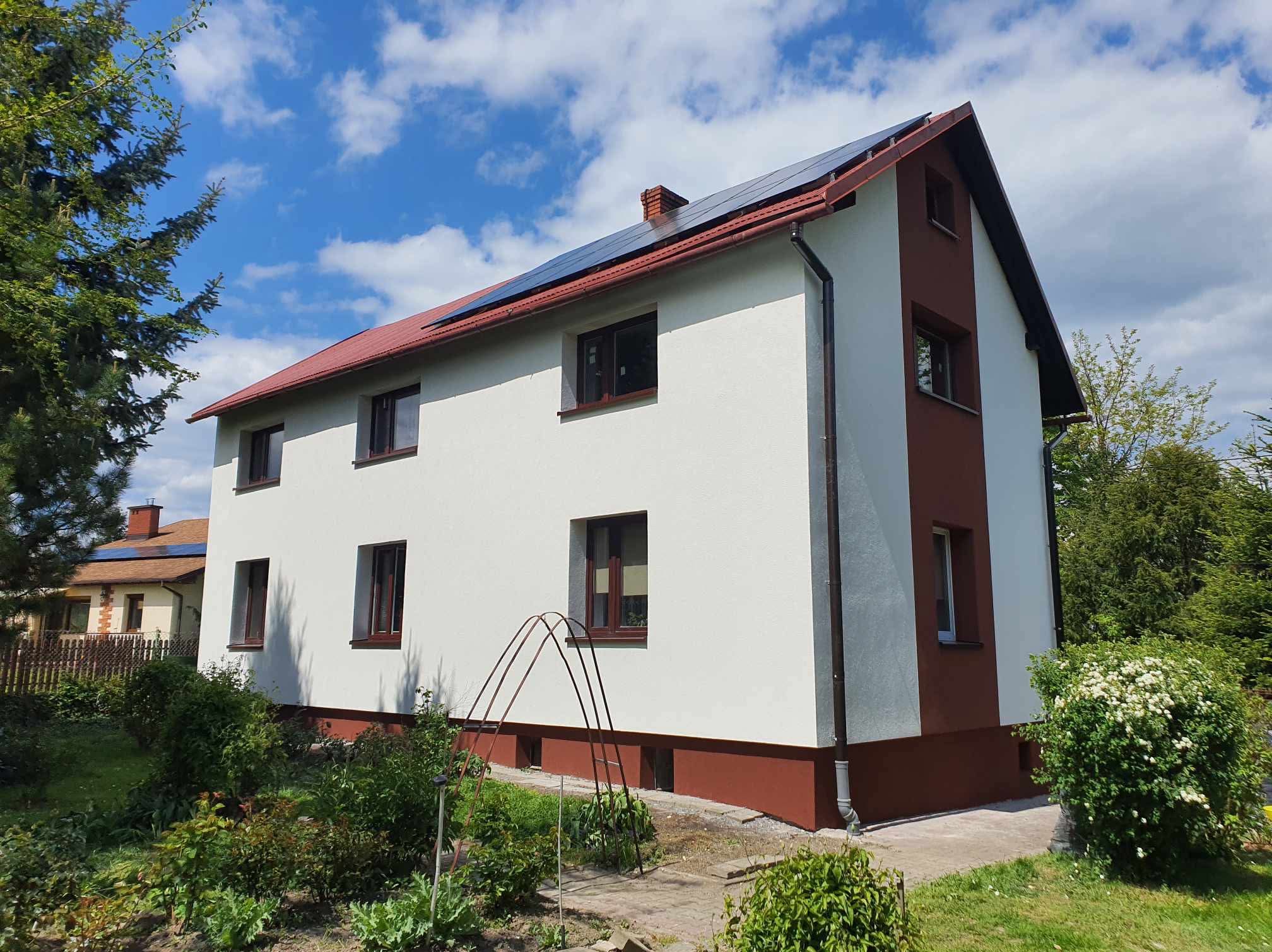 Dwukondygnacyjny dom z panelami słonecznymi na czerwonym dachu w Milówce. Biała elewacja z brązowymi akcentami. Widok z ogrodu w słoneczny dzień.