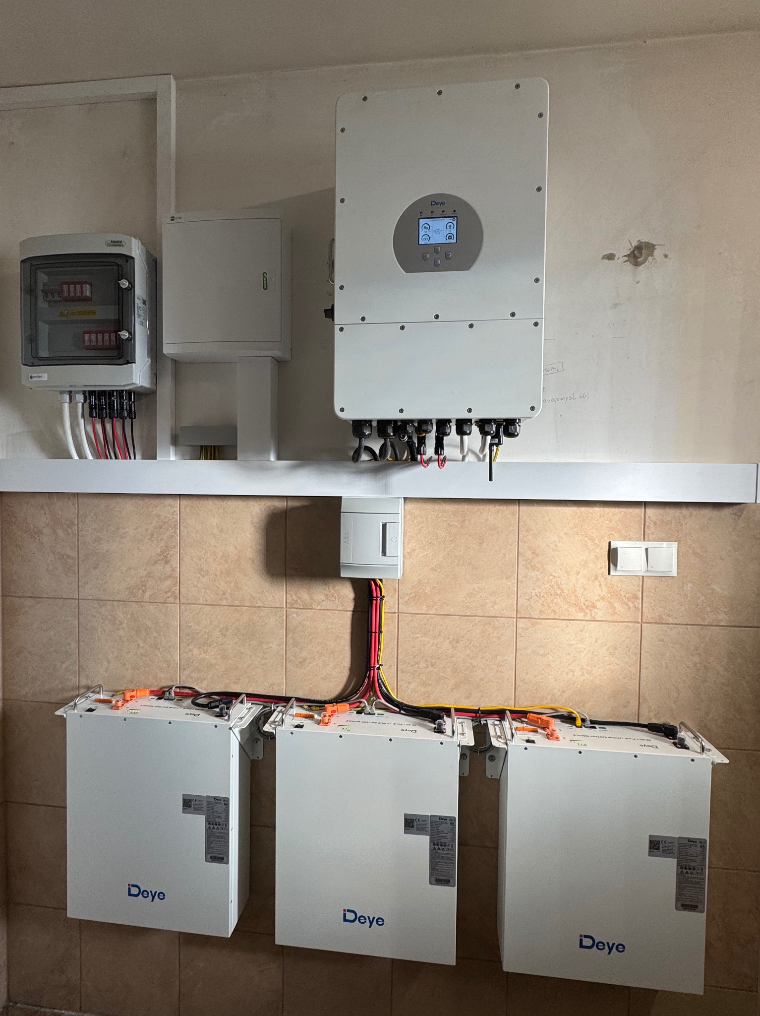 Instalacja magazynu energii Deye: trzy białe moduły na ścianie z brązowymi płytkami, inwerter i rozdzielnia elektryczna. Widoczne przewody i złącza.