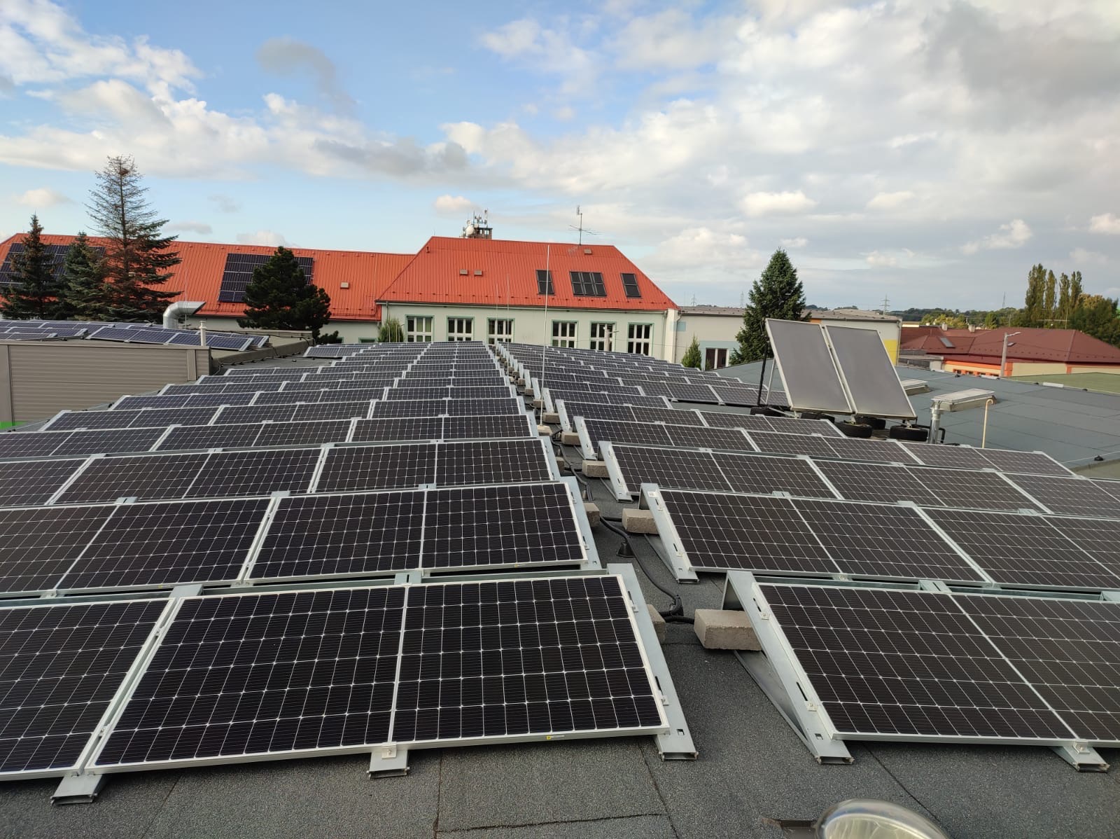 Rozległa instalacja paneli słonecznych na dachu budynku z czerwoną dachówką w tle, widok z góry, pochmurne niebo. Układ paneli w rzędach, widoczne mocowania.