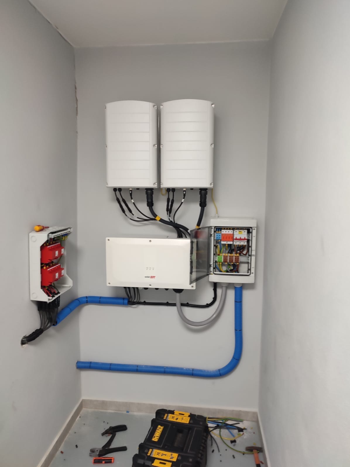 Zestawienie urządzeń: dwa inwertery fotowoltaiczne, skrzynka z bezpiecznikami i rozdzielnica, połączone przewodami w peszlach na szarej ścianie. Widoczne narzędzia.