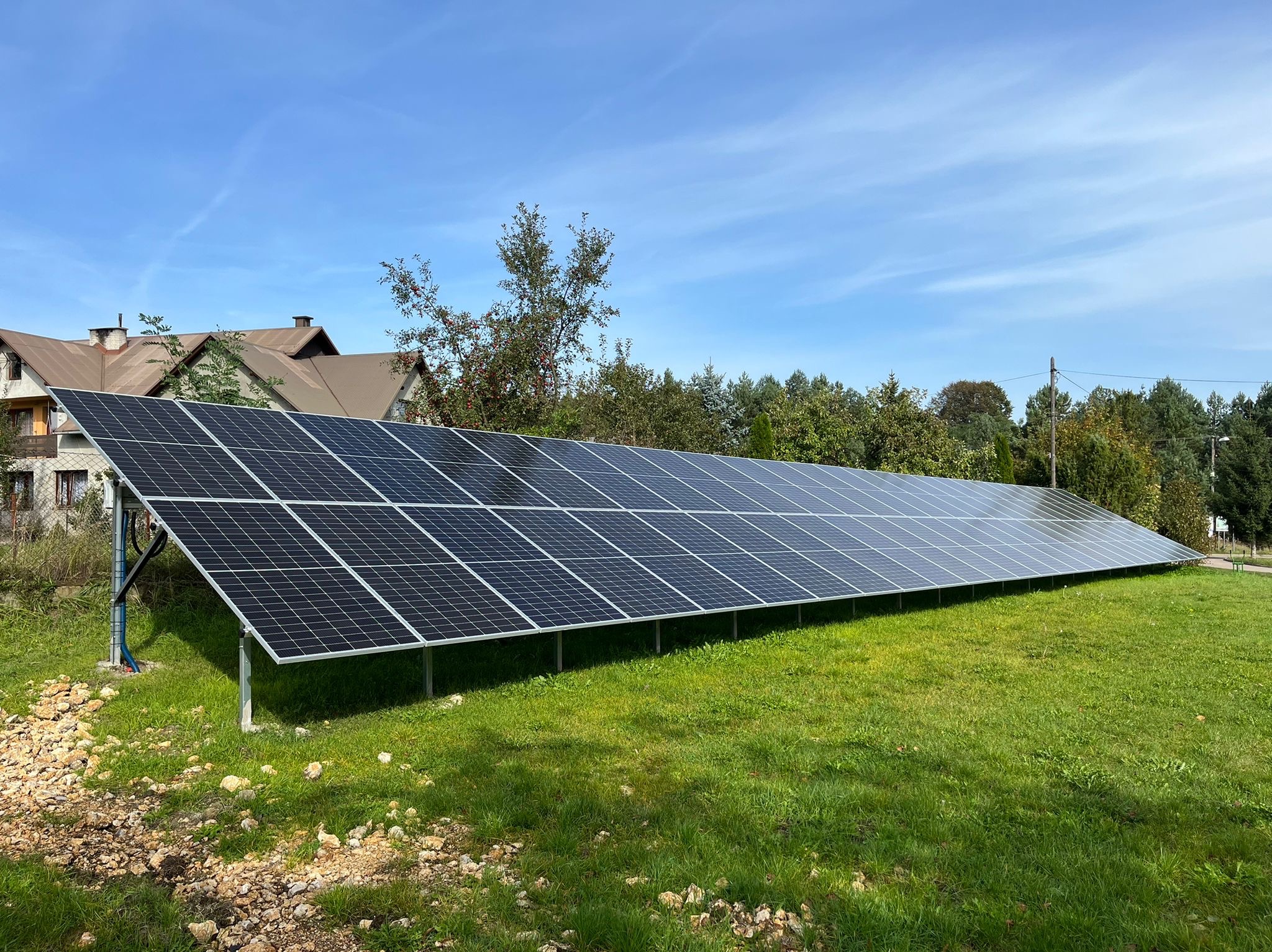Długi rząd paneli fotowoltaicznych na zielonej trawie, z domem w tle pod błękitnym niebem. Instalacja na gruncie, widoczne mocowania i kable.
