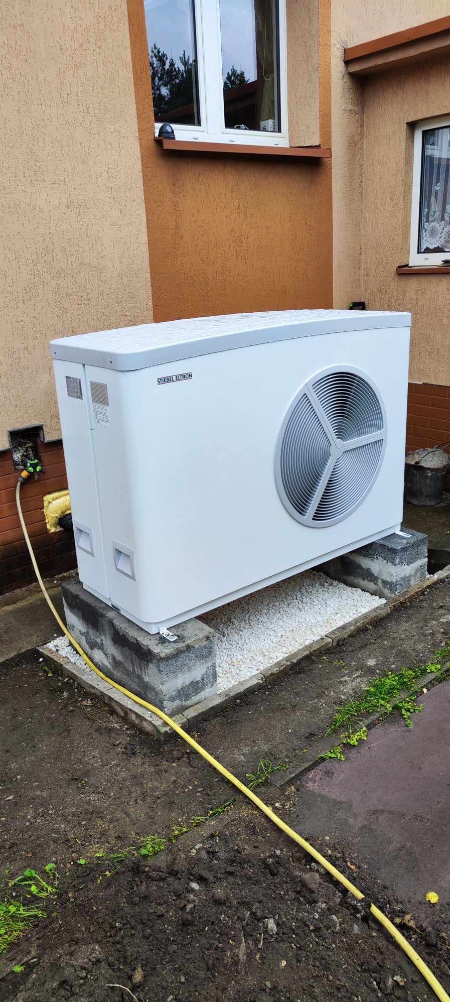 Biała pompa ciepła Stiebel Eltron na betonowych blokach i białym żwirze przed budynkiem z oknami, widoczny fragment żółtego węża.