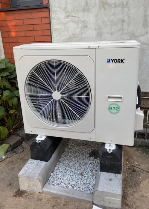 Biała jednostka zewnętrzna pompy ciepła YORK, umieszczona na betonowym fundamencie z szarym żwirem, na tle ściany budynku z cegły i betonu.