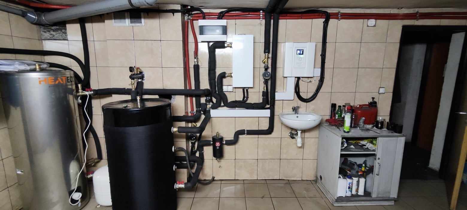 Złożona instalacja grzewcza w kotłowni: zbiorniki, rury z izolacją, sterownik. Widoczne elementy instalacji wodnej i kanalizacyjnej. Tło stanowią kafelki.