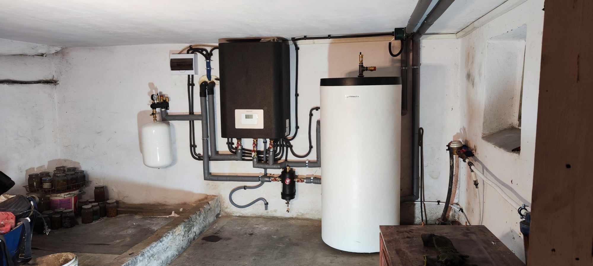 Zestaw kotłowni w piwnicy: czarny kocioł z wyświetlaczem, biały zbiornik akumulacyjny, białe naczynie przeponowe, orurowanie. Widoczne liczniki i zawory.