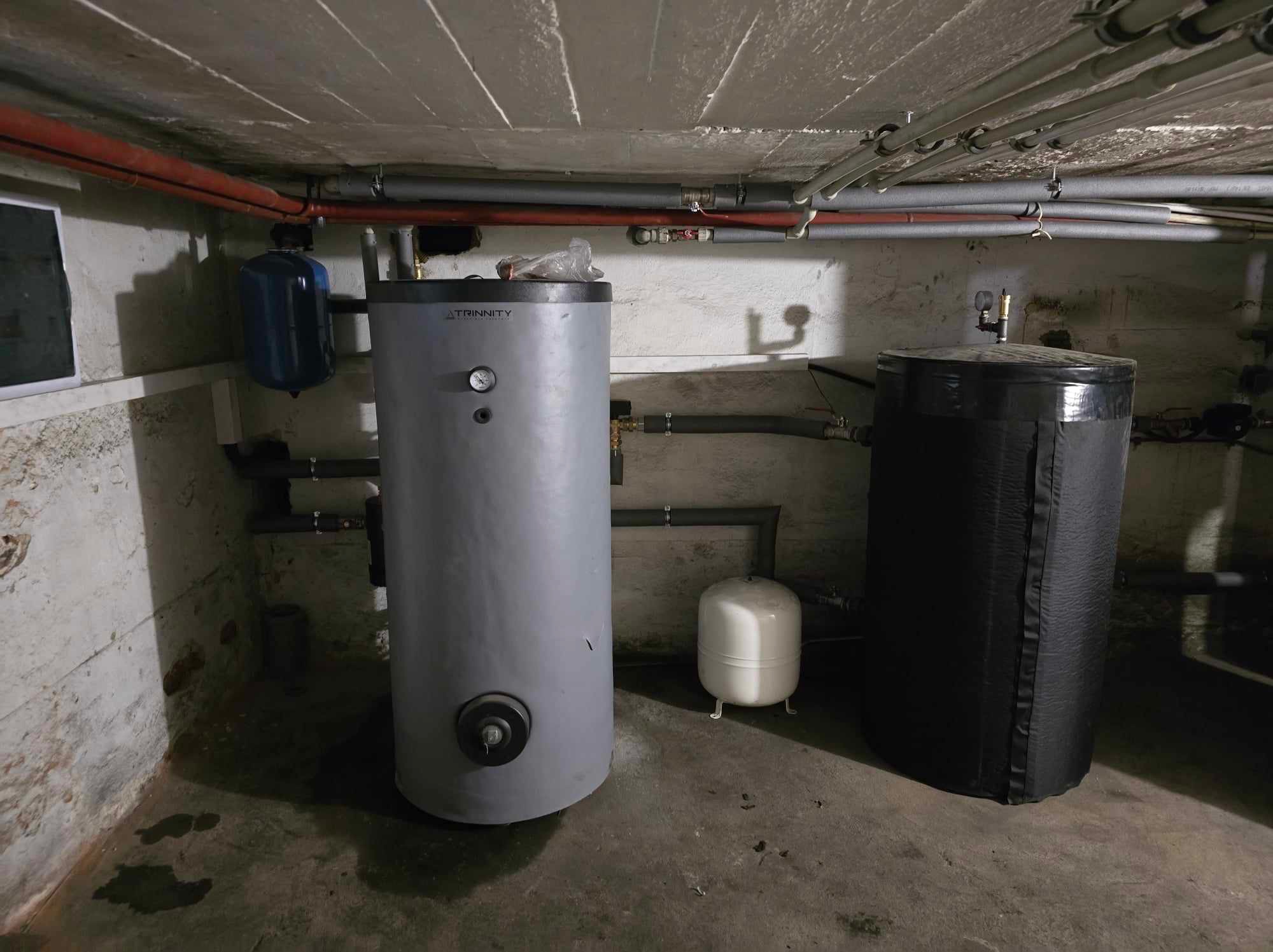 Szary zbiornik Trinnity z manometrem i czarny zbiornik w piwnicy, widoczne rury i małe zbiorniki ciśnieniowe. Instalacja w surowym wnętrzu.