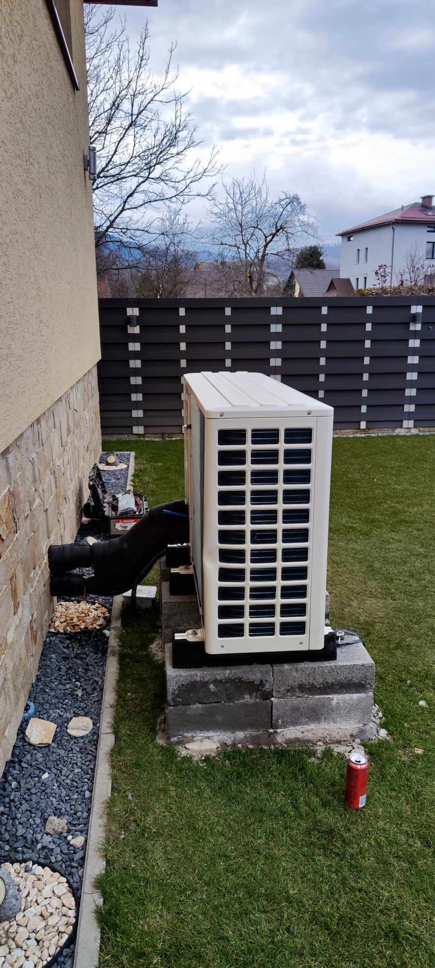 Biała pompa ciepła na betonowej podstawie przy ścianie domu. Widoczne czarne rury, trawnik i puszka po napoju. W tle ogrodzenie i budynki.