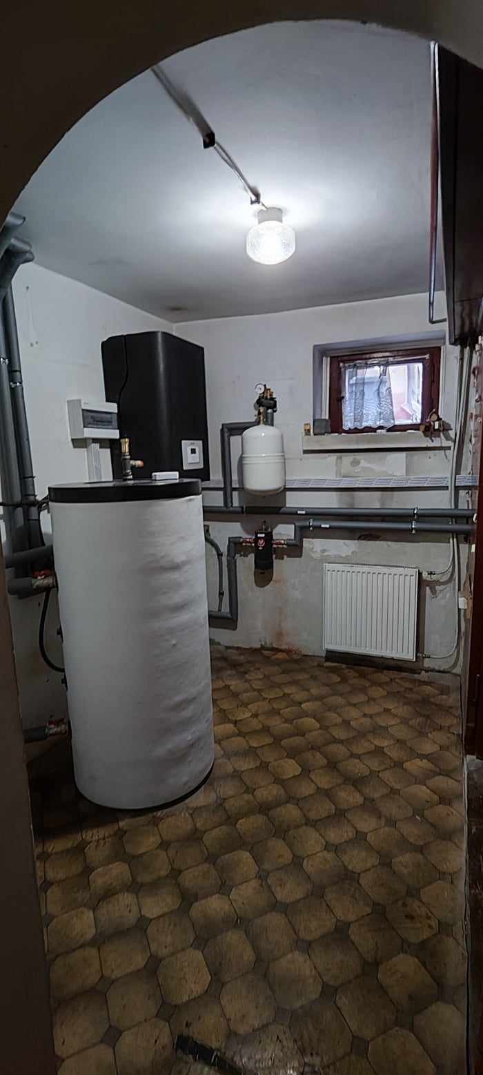 Kotłownia z białym zasobnikiem ciepłej wody i czarnym zbiornikiem buforowym. Widoczne rury instalacji grzewczej, grzejnik i okno.