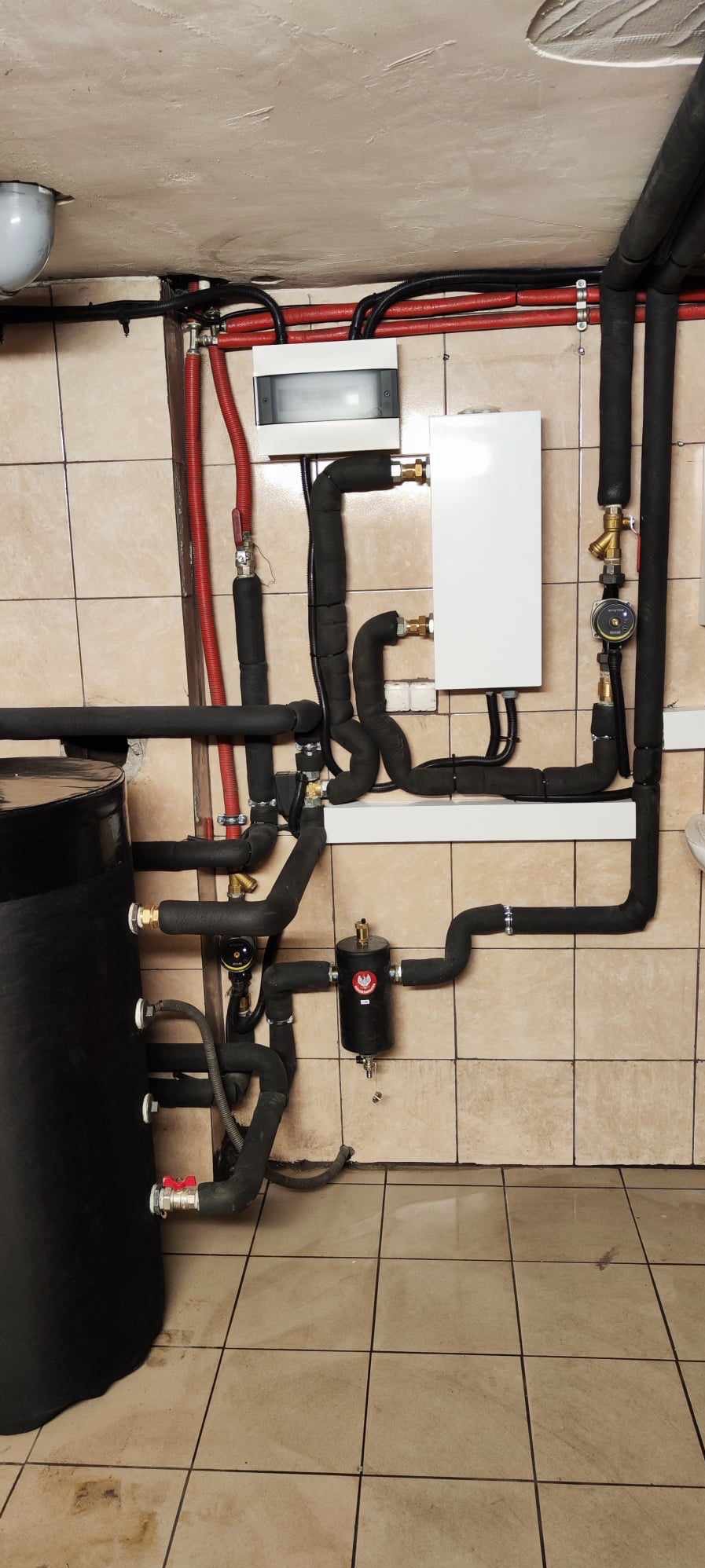 Złożona instalacja grzewcza z czarnym zbiornikiem, białym piecem i rurami w izolacji. Widoczne liczne zawory, manometry i elementy łączeniowe. Tło stanowią kafelki.