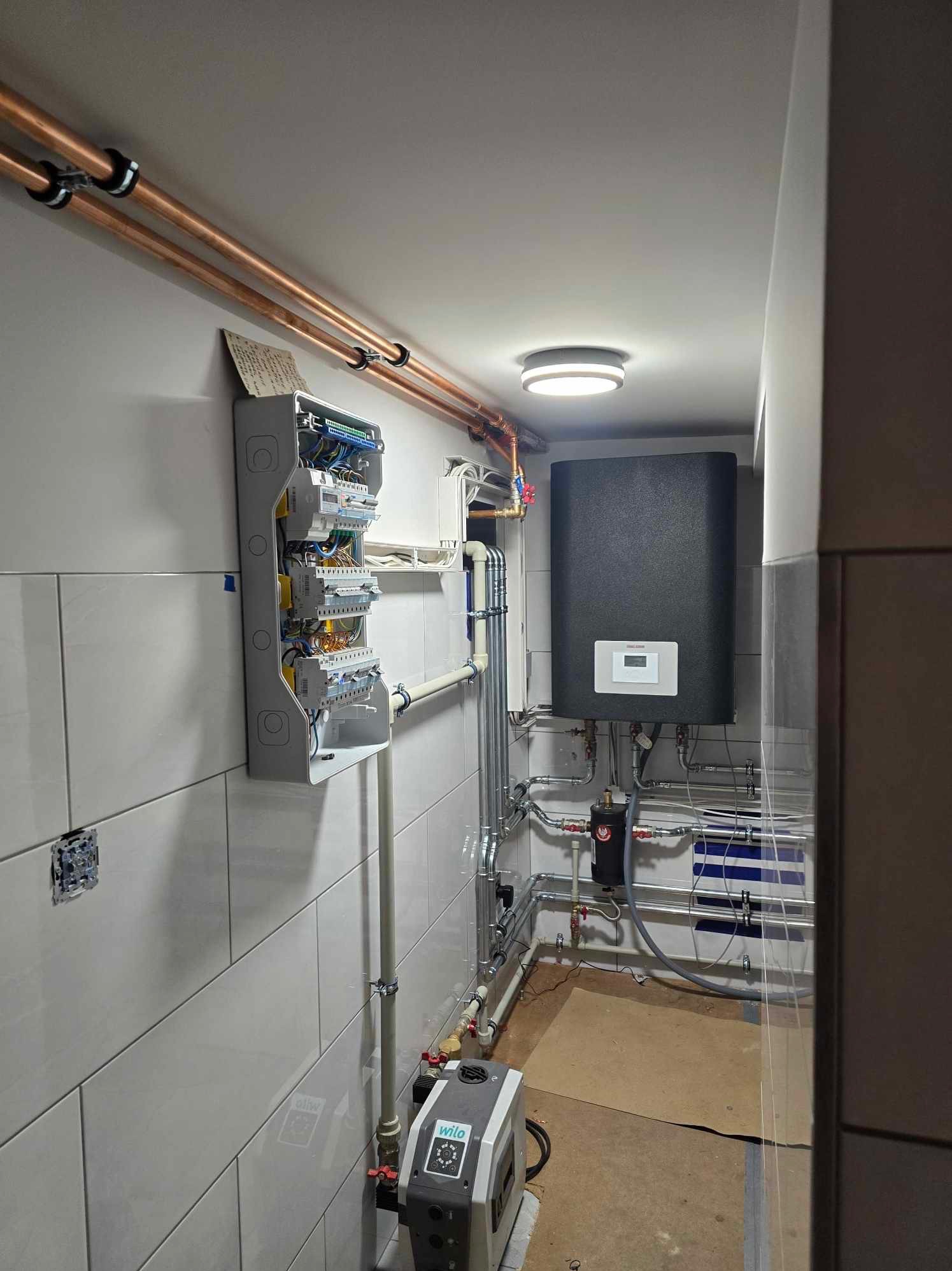 Węzeł cieplny w kotłowni: czarny zasobnik, rozdzielnia elektryczna z okablowaniem na kafelkowanej ścianie, rury miedziane pod sufitem, pompa obiegowa Wilo.