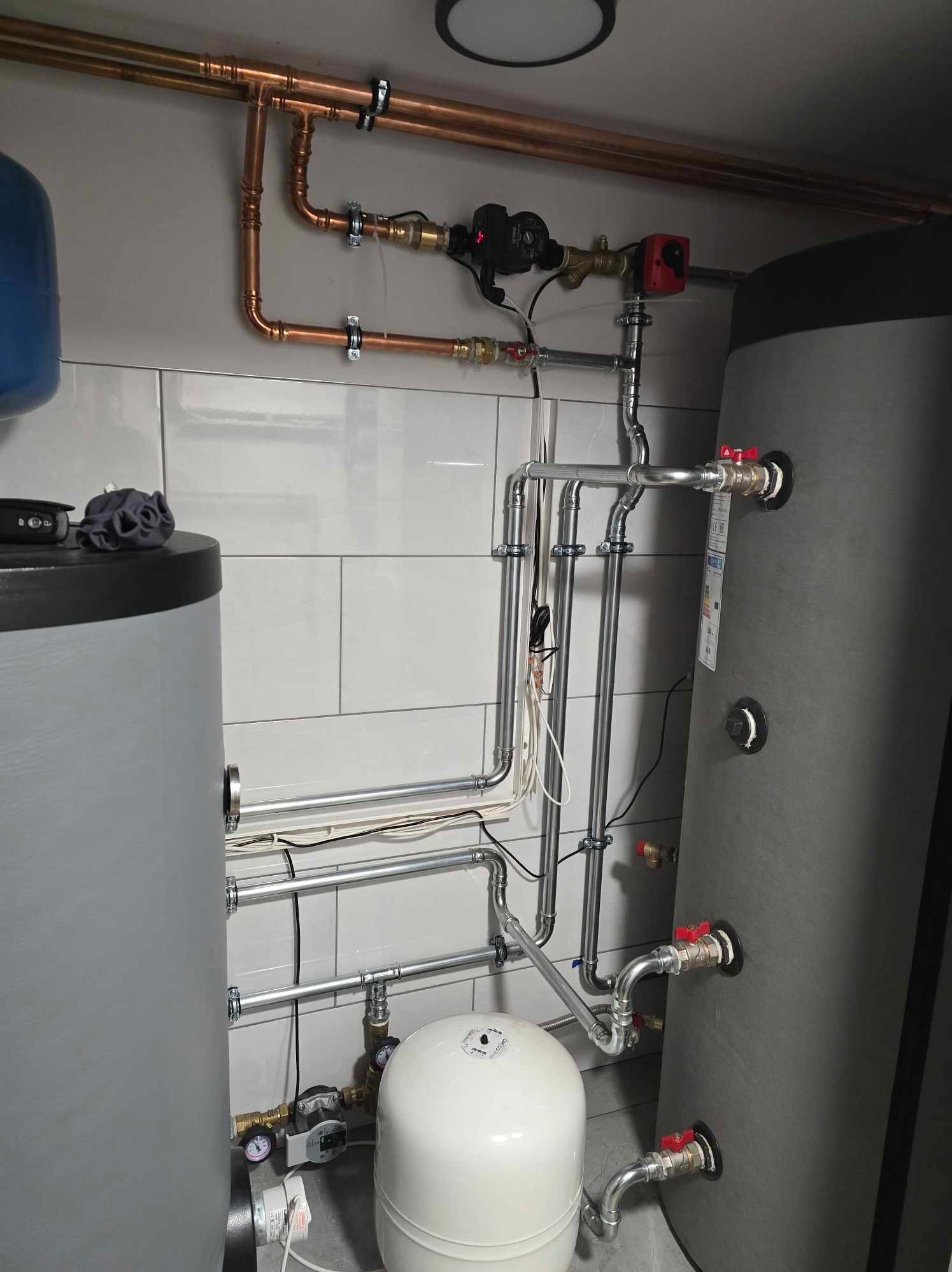 Instalacja rur miedzianych i stalowych z zaworami, manometrami i pompą obiegową w kotłowni z dwoma zbiornikami akumulacyjnymi, na tle białych płytek.