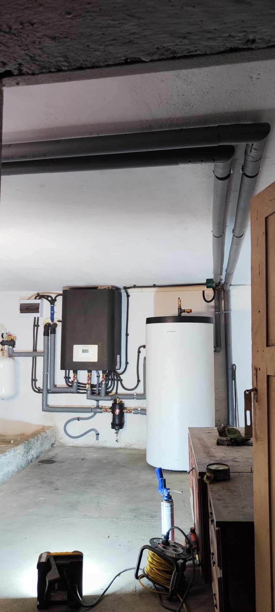 Zainstalowana instalacja z zbiornikiem i czarnym panelem na ścianie, liczne rury, widoczne podłączenia. Elementy instalacji w piwnicy.