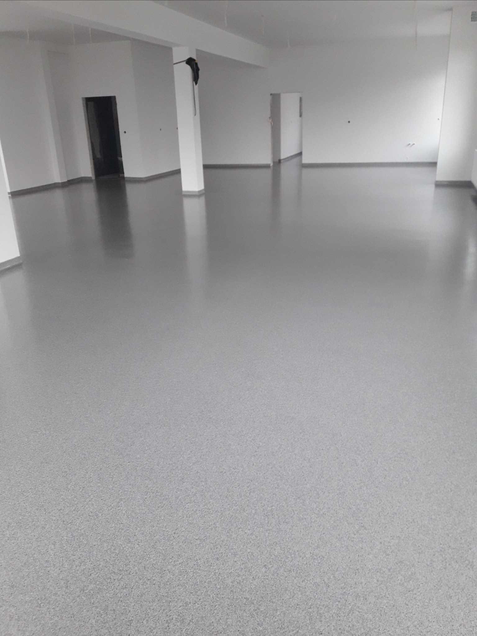 Szara, gładka powierzchnia posadzki w przestronnym, jasnym pomieszczeniu z białymi ścianami i filarami. Widoczne refleksy światła na podłodze. Elementem wyróżniającym jest ubranie wiszące na filarze.