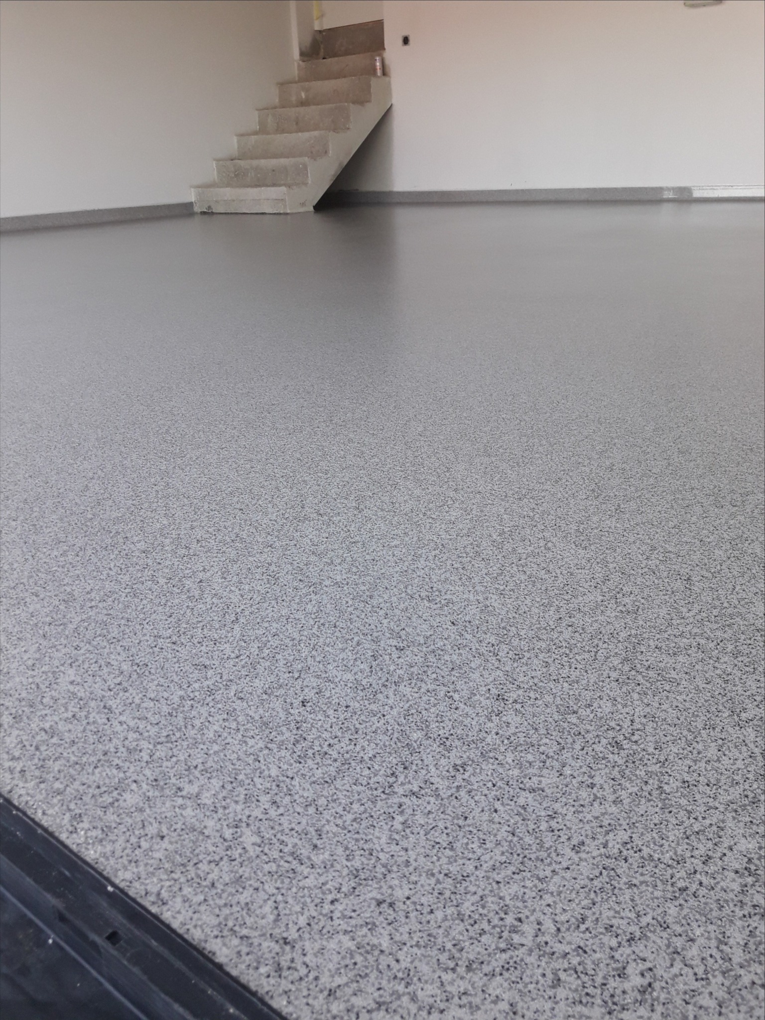 Szara, wzorzysta podłoga w hali przemysłowej, z betonowymi schodami w tle i widocznymi fragmentami czarnej listwy przypodłogowej w lewym dolnym rogu kadru.