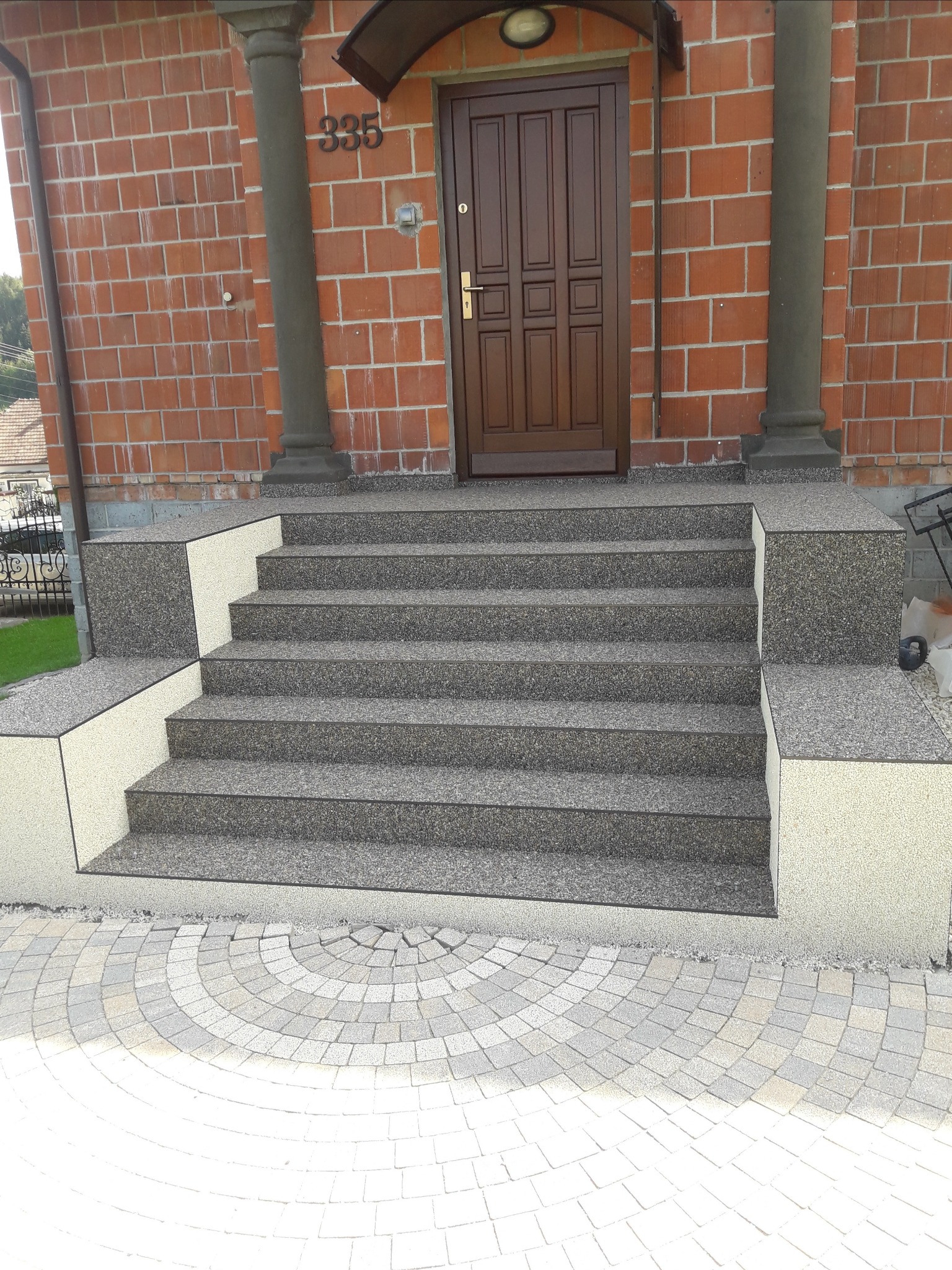 Schody wejściowe wykończone szarą, kamienną posadzką z żywicą, prowadzące do brązowych drzwi wejściowych z numerem 335, na tle ceglanej ściany z kolumnami.