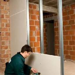 Pracownik w zielonym kombinezonie przykręca płytę gipsowo-kartonową do metalowego stelaża w surowym wnętrzu z ceglanymi ścianami.