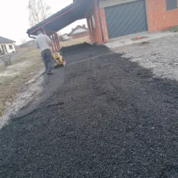 Pracownik zagęszcza świeżo rozłożoną, ciemną nawierzchnię drogi w pobliżu budynku z garażem i zadaszeniem dla samochodu.