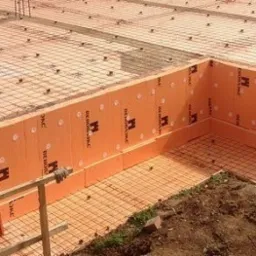 Pomarańczowe płyty izolacyjne z logo producenta ułożone na fundamencie budynku, zabezpieczone siatką zbrojeniową i plastikowymi kołkami.