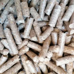 Stos brązowo-szarych, cylindrycznych brykietów z widoczną strukturą włókien drzewnych, ułożonych chaotycznie.