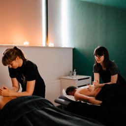 M&M Zdrowy Masaż - Dwie terapeutki wykonują zabiegi na plecach klientów w nastrojowym gabinecie z ciepłym oświetleniem świec i ścianami w odcieniach beżu i ciemnej zieleni.