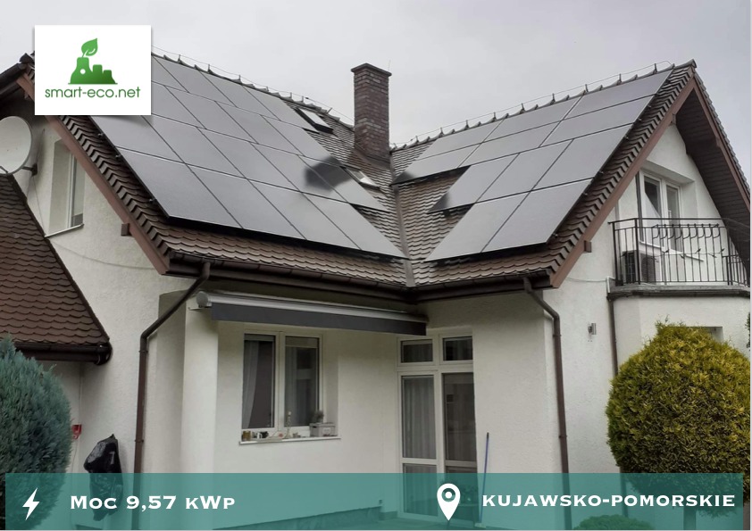 Dom jednorodzinny z panelami słonecznymi na dachu, widoczny komin i okna dachowe, z przodu budynku krzewy ozdobne i fragment okna z roletą.