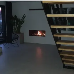 Nowoczesny salon z minimalistycznym biokominkiem w ścianie, drewnianymi schodami z czarną metalową konstrukcją i widokiem na ogród przez duże okno.