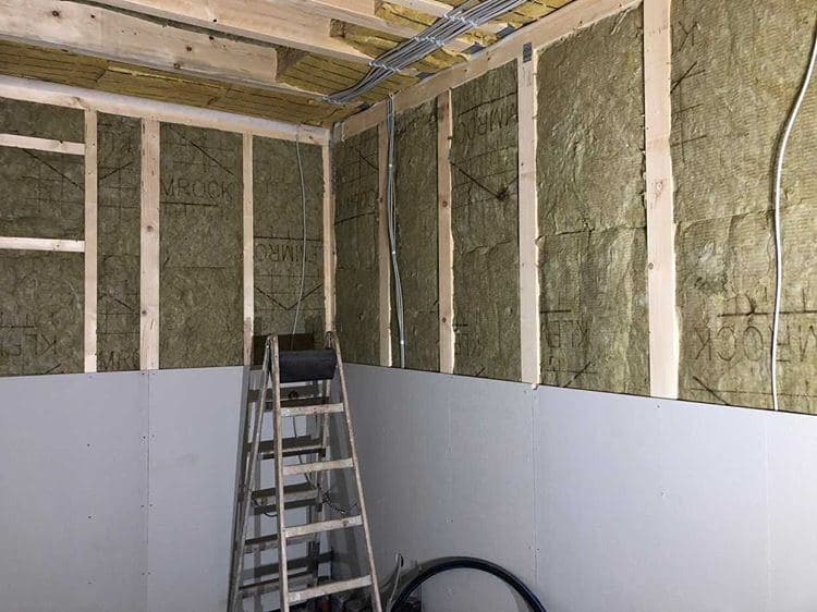 Wnętrze w trakcie prac ociepleniowych: widoczne drewniane legary, wełna mineralna z nadrukiem 'MROCK', częściowo zamontowane płyty gipsowo-kartonowe oraz aluminiowa drabina oparta o ścianę.