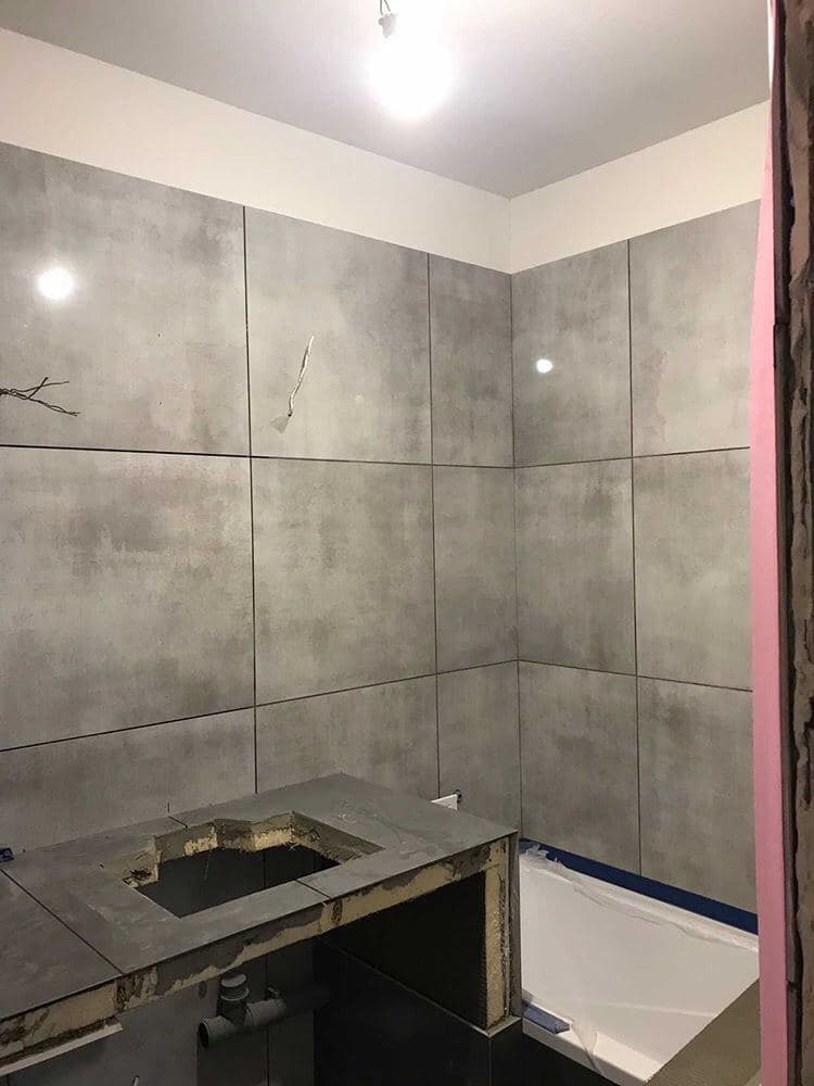 Łazienka w trakcie remontu: szare, matowe płytki wielkoformatowe na ścianach, przygotowany blat pod umywalkę z wyciętym otworem, widoczna wanna zabezpieczona folią, surowy stan wykończenia.