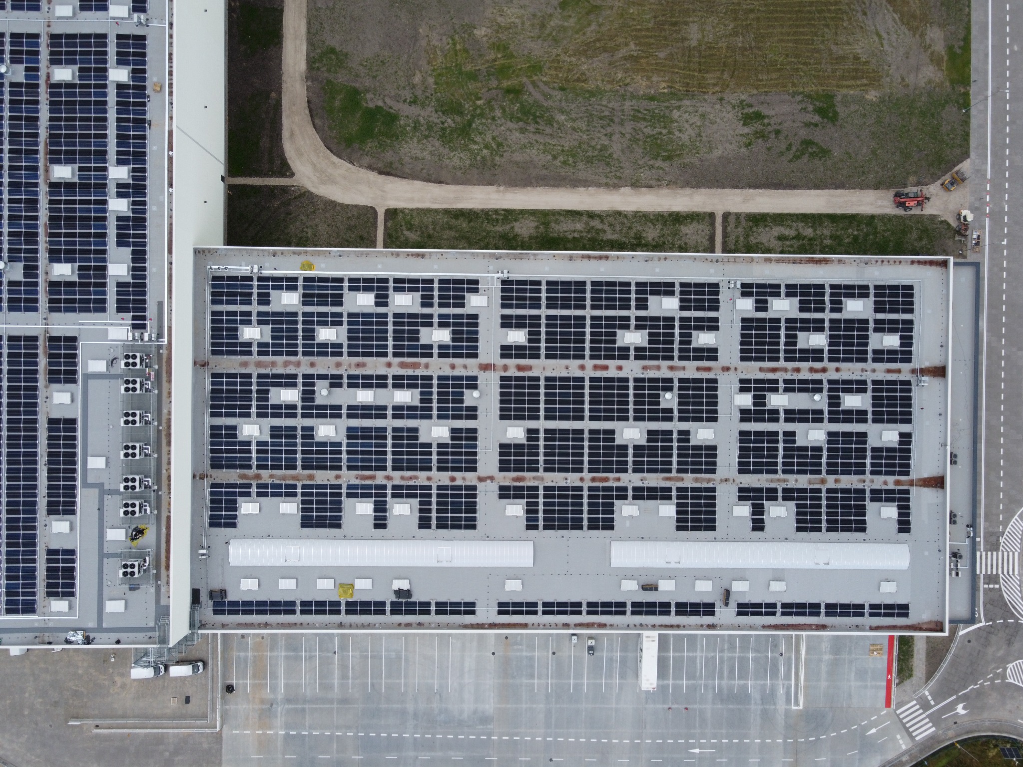 Widok z góry na rozległą instalację paneli słonecznych na dachach budynków przemysłowych, z widocznym parkingiem i fragmentem terenu zielonego w tle.