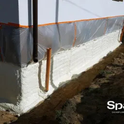 Izolacja fundamentów pianką PUR z zabezpieczeniem folią i taśmą, widoczna rura spustowa i fragment elewacji budynku.