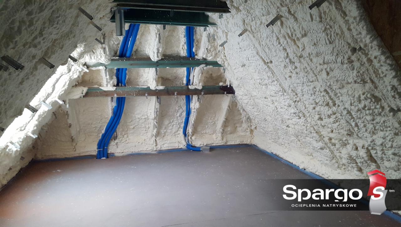 Poddasze z widoczną drewnianą konstrukcją i niebieskimi rurami, pokryte grubą warstwą białej pianki izolacyjnej, z widocznym logo firmy na podłodze.