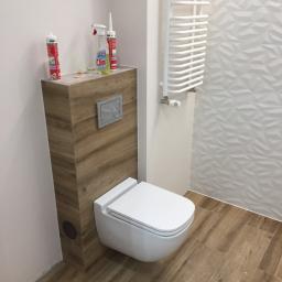 Łukasz Budownictwo - Wisząca toaleta z zabudową w odcieniach drewna, biały grzejnik drabinkowy i trójwymiarowa struktura płytek na ścianie, widoczne materiały budowlane na zabudowie i podłodze.